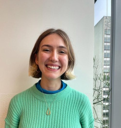 Ellie is wearing a green jumper and has short blonde hair, and is smiling against a white wall